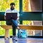 Student sitting on a bench in front of a large window with their laptop open on their lap. 