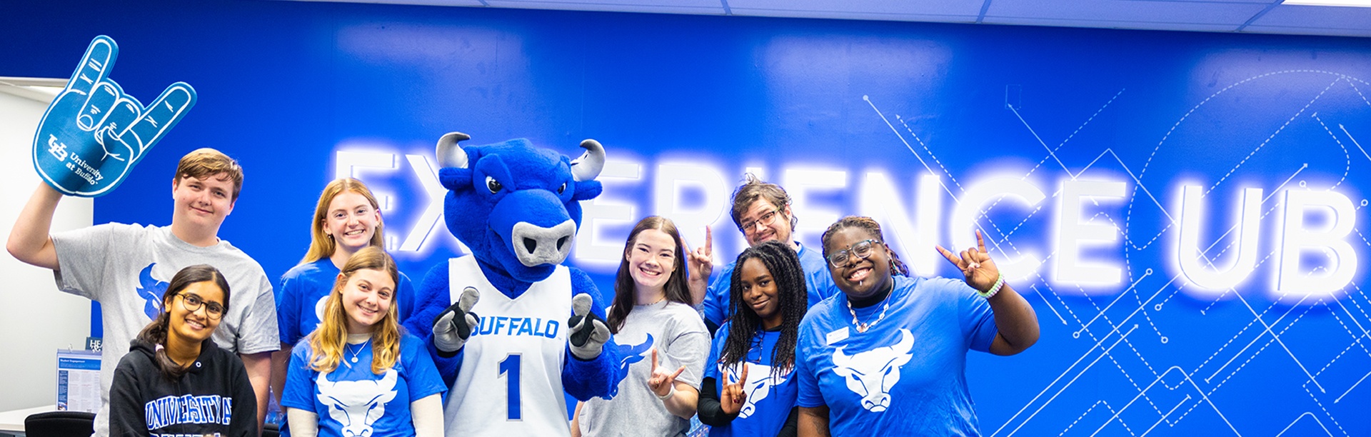 Students pose together in front of a wall reading &ldquo;Experience UB.&rdquo;. 