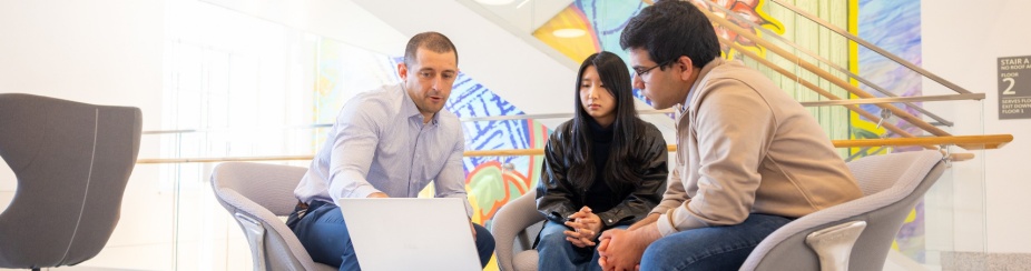 Two students and a professor sitting around a table looking at a laptop together. 