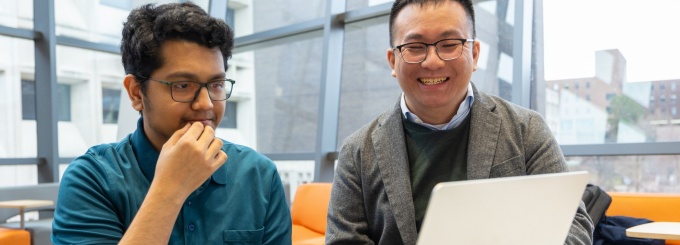 Student and faculty member are sitting side by side looking at a laptop together. 